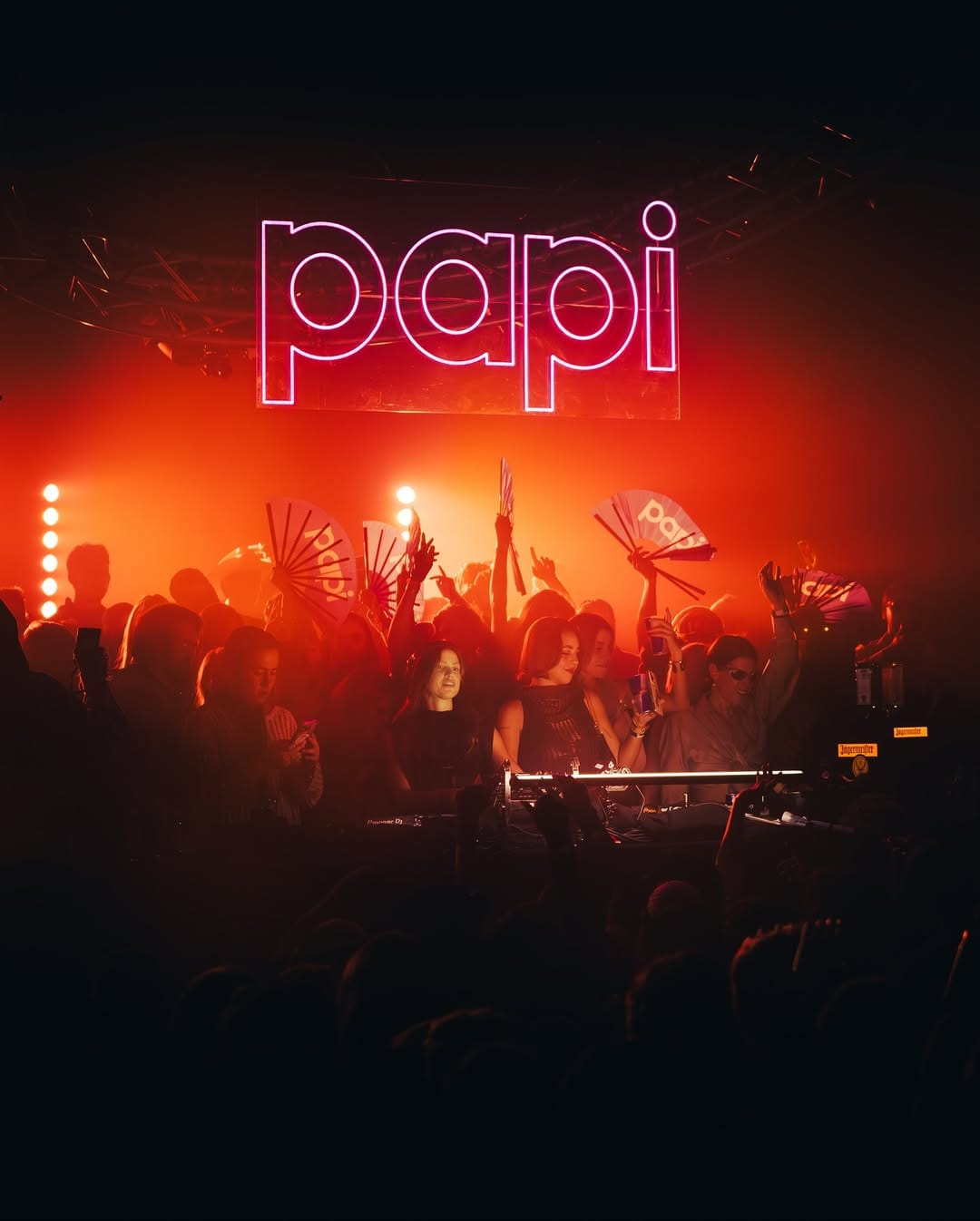 Neon sign with 'papi' over a crowd at a concert with red lighting.